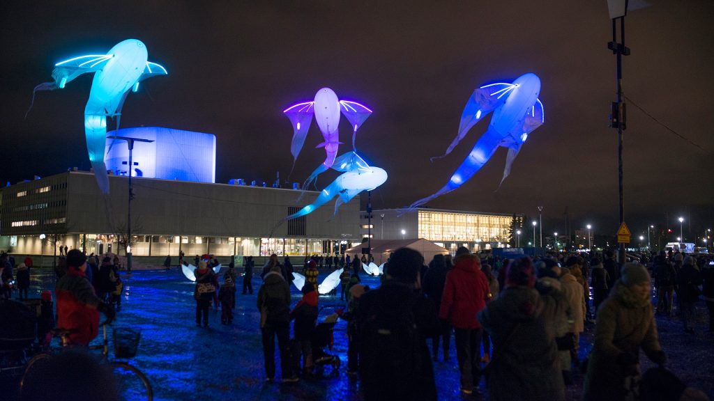 A bluish and purple-toned light installation in Oulu's market square, people standing near the installation, Oulu Theater in the background