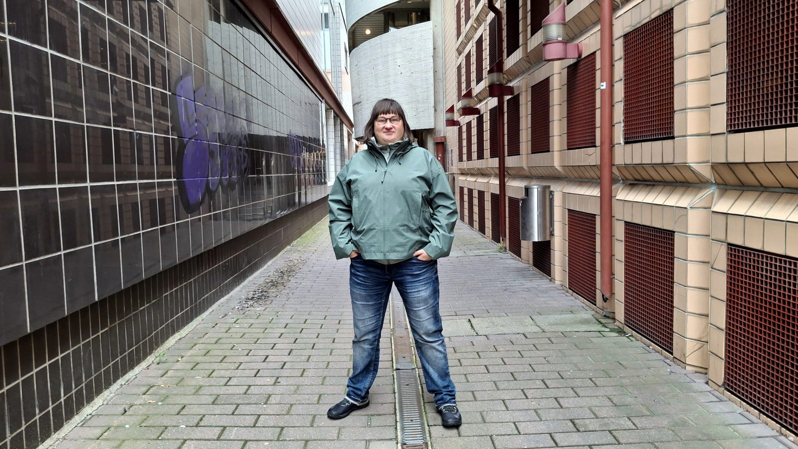 Nainen seisoo käytävällä vihreässä takissa ja farkuissa